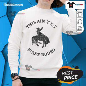 Alt Text: Cowboy wearing an Ain't My First Rodeo sweatshirt, riding a bucking bronc horse at a rodeo event.