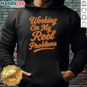 Person wearing a 'Happy Working On My Reel Problems' fishing hoodie, smiling while holding fishing gear outdoors.