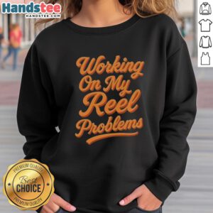 Person wearing a 'Happy Working On My Reel Problems' fishing sweatshirt, smiling outdoors by a serene lake.